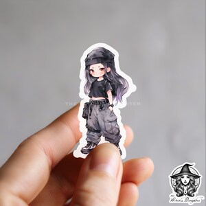 Puede incluir: Una pegatina con una ilustraci&oacute;n de dibujos animados de una persona joven con cabello negro que lleva una boina negra, un top corto negro y pantalones cargo grises. El personaje lleva una bolsa negra con una correa blanca.