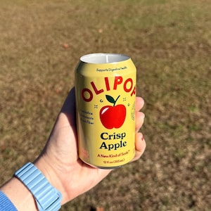 May include: A yellow can with the text "OLIPOP Crisp Apple" and a red apple graphic. The can is held in a hand with a blue watch. The can is 12 fl oz (355 mL).