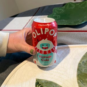 May include: A red and white Olipop Vintage Cola can candle with a smiling yeti graphic. The can is 12 fl oz (355ml). The candle is on a table with a green leaf-shaped plate.
