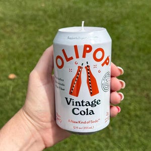 May include: A white candle shaped like a soda can, with the text "OLIPOP Vintage Cola" in orange. The can design includes orange bottles with straws. The candle is 12 fl oz (355mL) and has a wick on top.