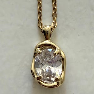 May include: A gold-colored necklace with a pendant featuring a large, clear gemstone. The pendant is irregularly shaped and set in a gold bezel. The chain is a delicate link style.