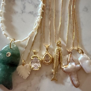 May include: A collection of gold necklaces with various pendants, including a green fish, a white heart, a clear oval stone, a teardrop-shaped stone, a fish, a cross, and a pearl. The necklaces are displayed on a light-colored surface.