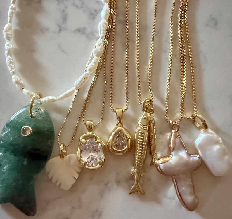 May include: A collection of gold necklaces with various pendants, including a green fish, a white heart, a clear oval stone, a teardrop-shaped stone, a fish, a cross, and a pearl. The necklaces are displayed on a light-colored surface.
