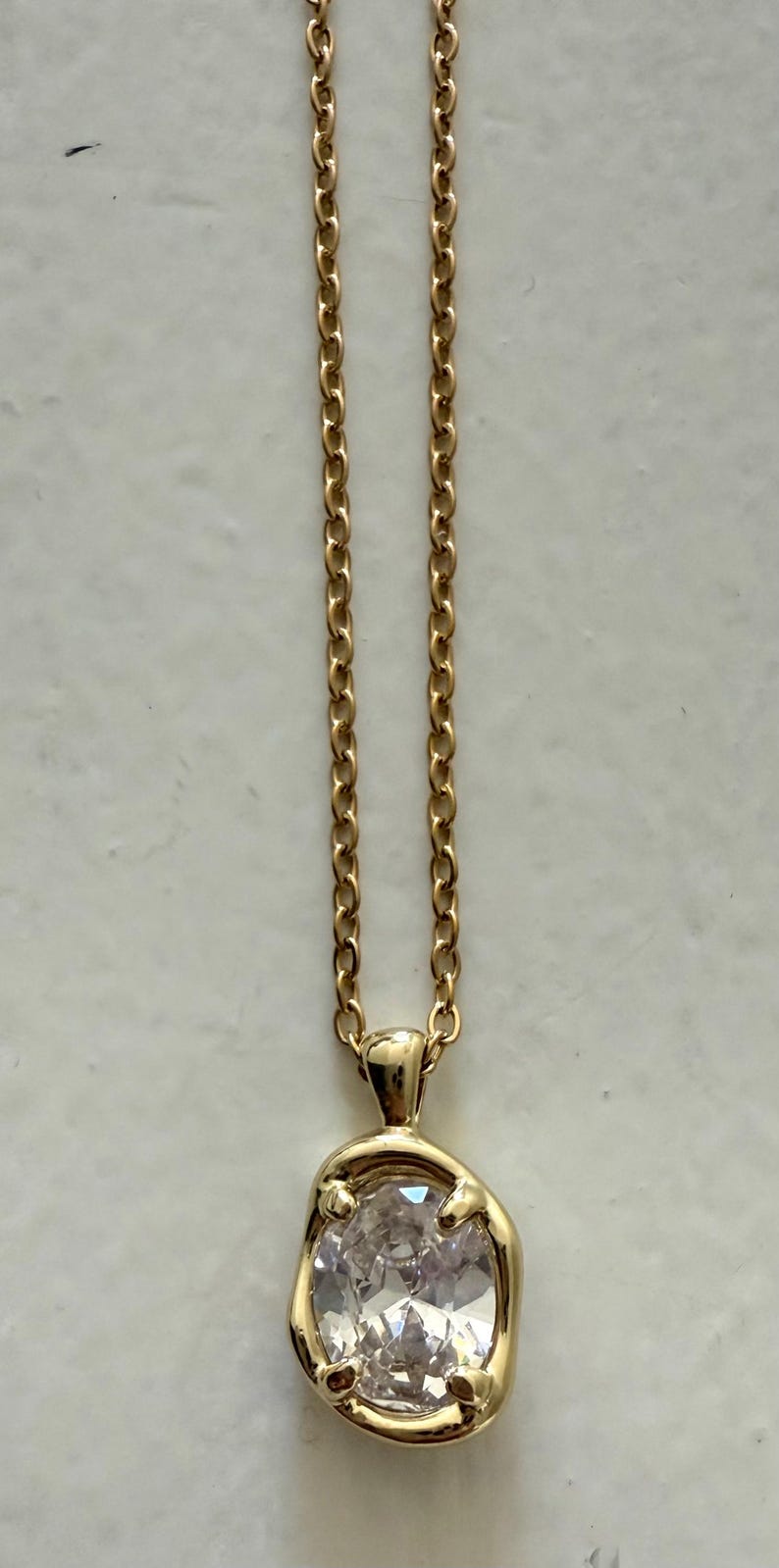 May include: A gold-colored necklace with a pendant featuring a large, clear gemstone. The pendant is irregularly shaped and set in a gold bezel. The chain is a delicate link style.
