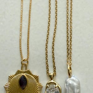 May include: Three gold necklaces with different pendants. One has a sunburst design with a dark purple stone, another has a clear, oval-shaped stone, and the third has a white, oblong pearl.