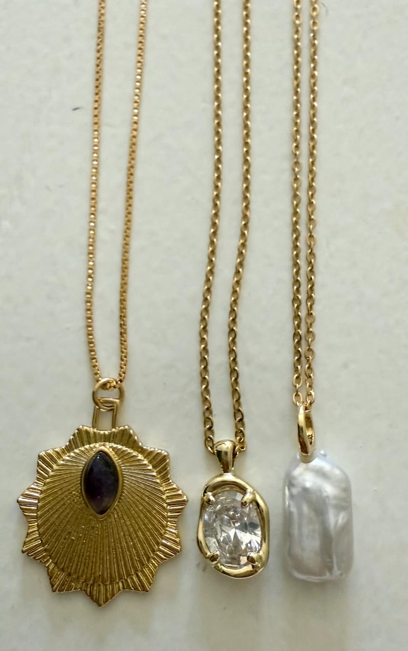 May include: Three gold necklaces with different pendants. One has a sunburst design with a dark purple stone, another has a clear, oval-shaped stone, and the third has a white, oblong pearl.