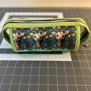 May include: A rectangular pencil case with a zipper closure. The case features a repeating pattern of giraffe faces on a teal background with orange flowers. The top and sides are green with white polka dots. The zipper is black and green.