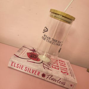 May include: A clear glass tumbler with a bamboo lid and a straw. The tumbler has the text "Wear the hat, ride the cowboy" printed on it. The tumbler is sitting on top of a book titled "Flawless" by Elsie Silver.
