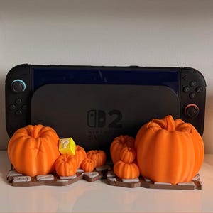 May include: A black gaming console dock featuring a Nintendo Switch logo, displayed with a collection of orange 3D-printed pumpkins of varying sizes arranged on a brown, textured base that resembles a stone path or brick surface.