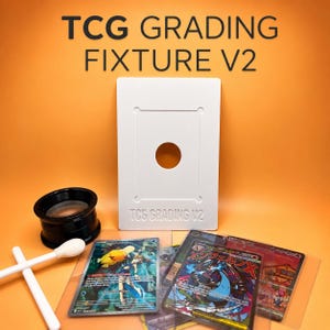 May include: A white TCG GRADING FIXTURE V2 rectangular tool with a center hole, a black magnifying loupe, two white cotton swabs, and three trading cards in clear plastic sleeves. The text TCG GRADING FIXTURE V2 is printed at the top.