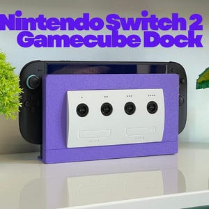 May include: A Nintendo Switch 2 Gamecube Dock in purple with a white Gamecube controller port design. The dock is designed to hold a Nintendo Switch console. The image also includes a green plant and a Mario-themed decoration.