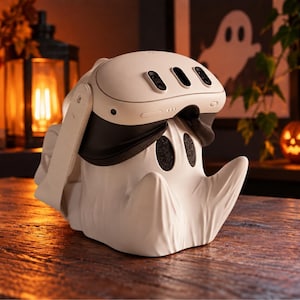 May include: A white VR headset stand shaped like a ghost with a smooth, matte finish. The stand features two dark, oval-shaped eye cutouts and a draped, sheet-like appearance. A white VR headset is resting securely on top of the ghost-shaped stand.