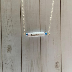 Puede incluir: Un collar de cadena plateada con un dije de rollo blanco que dice "Lucas 6:38". El dije está acentuado con dos pequeñas cuentas azules.
