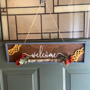 May include: A rectangular wooden welcome sign with a blue frame, brown center, and decorative yellow cutouts. The word "welcome" is written in white script. Orange flowers and a burlap ribbon add detail.