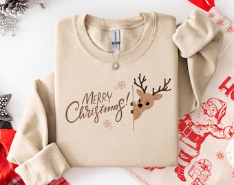 May include: A beige crewneck sweatshirt with the words "Merry Christmas!" in brown script, accompanied by a cartoon reindeer peeking from behind a line. The sweatshirt is folded, with a tag visible at the neckline. A festive holiday theme.