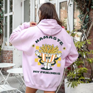 Könnte beinhalten: Hellrosa Kapuzenpullover mit dem Wort "NAMASTE" darüber. Darunter befindet sich ein Cartoon-Bild eines Behälters mit Pommes Frites mit Armen und Beinen. Der Text "MY FRIE(ND)S" ist darunter gedruckt.