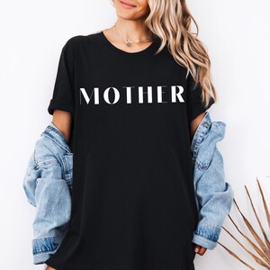 Könnte beinhalten: Schwarzes T-Shirt mit dem weißen Aufdruck "MOTHER" in einer fetten, serifenlosen Schriftart. Das T-Shirt wird mit einer hellblauen Jeansjacke getragen. Der Stil ist lässig und bequem.