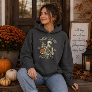 Könnte beinhalten: Ein dunkel anthrazitfarbener Kapuzenpullover mit einer Grafik eines Skeletts, das eine Topfpflanze hält. Der Text lautet DEAD PLANT SOCIETY mit EST. 2021. Der Hoodie verfügt über eine Kängurutasche, lange Ärmel und eine Kapuze mit Kordelzug.
