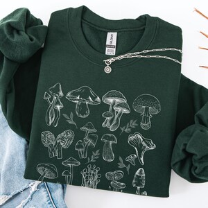 Peut inclure: Un sweat-shirt vert foncé avec une illustration blanche de divers champignons et feuilles. Un collier argenté avec un pendentif en forme de soleil est drapé sur le sweat-shirt. Le sweat-shirt est plié sur un jean bleu.