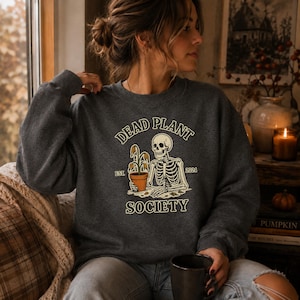 Könnte beinhalten: Ein dunkelgraues Sweatshirt mit Rundhalsausschnitt und einer Grafik eines Skeletts, das eine verwelkte Topfpflanze hält. Der Text lautet DEAD PLANT SOCIETY mit EST. 2024 auf beiden Seiten. Das Design verwendet Creme-, Orange- und Brauntöne.