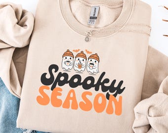 May include: A beige sweatshirt with the words "Spooky Season" in black and orange. Above the text are three cartoon ghosts wearing orange beanies and holding beverages. A silver necklace with a sun pendant is on the sweatshirt.