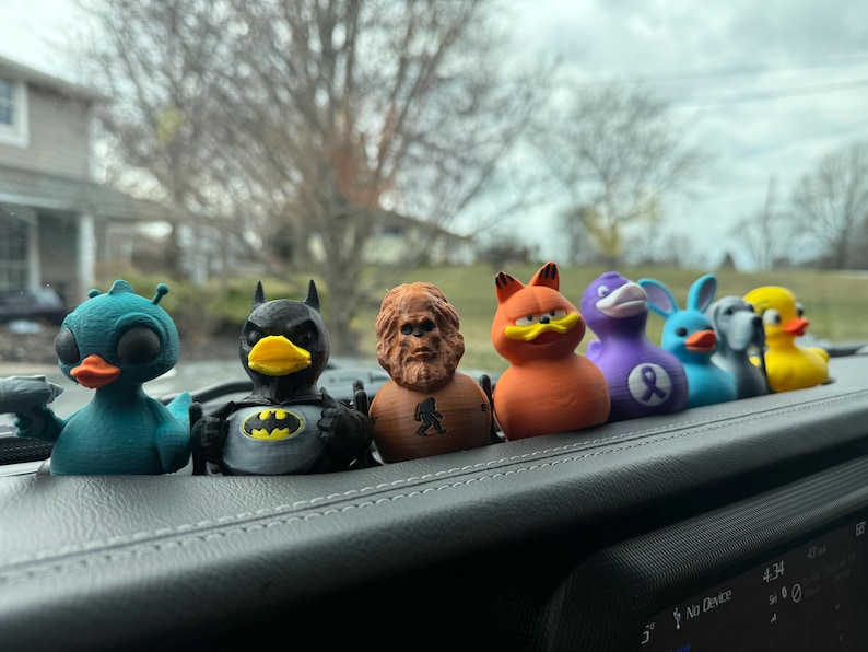 K&ouml;nnte beinhalten: Eine Reihe von sieben bunten Gummiente-Figuren, darunter eine blaue Ente mit einem au&szlig;erirdischen Kopf, eine schwarz-gelbe Batman-Ente, eine braune Bigfoot-Ente, eine orangefarbene Garfield-Ente, eine lila Ente mit einem Band, eine blaue Ente mit Hasenohren und eine grau-wei&szlig;e Ente. Die Enten sitzen auf einem schwarzen Armaturenbrett.