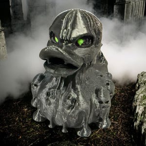 May include: A black rubber duck with green eyes, dripping with a melted, gooey texture. The duck is in a spooky setting with fog and stone structures.