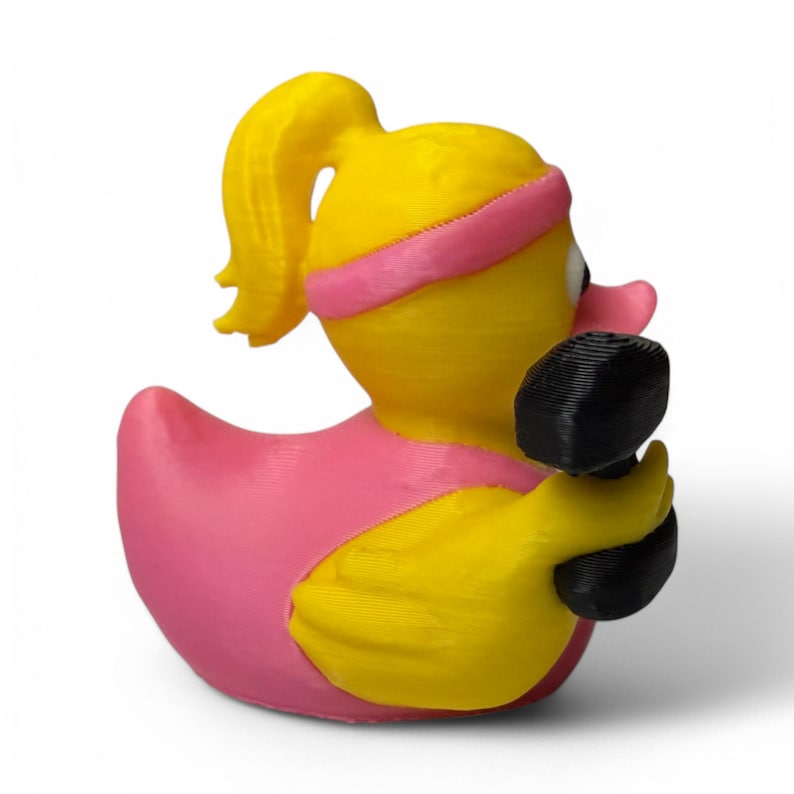 K&ouml;nnte beinhalten: Eine gelbe Gummiente mit einem rosafarbenen Stirnband und einer schwarzen Hantel in jeder Hand. Die Ente hat einen gelben Schnabel und ein schwarzes Auge.