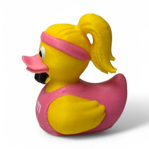 K&ouml;nnte beinhalten: Eine gelbe Gummiente mit einem rosafarbenen Sport-BH und einem Stirnband. Die Ente hat einen Pferdeschwanz und ein schwarzes Auge.