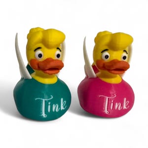 May include: Two yellow rubber duckies with white wings. One duck is wearing a teal blue shirt with the word "Tink" written on it. The other duck is wearing a pink shirt with the word "Tink" written on it.