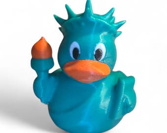May include: A teal-colored duck figurine with an orange beak and torch. The duck has a spiky crown, resembling the Statue of Liberty. The figurine is a novelty item, likely a collectible or decorative piece.