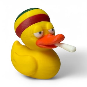 May include: A yellow rubber duck with an orange beak and a white object in its mouth. The duck wears a headband with green, yellow, and red stripes. The duck has a slightly grumpy expression.