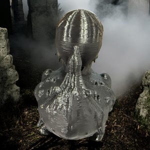 May include: A 3D printed sculpture of a creature with a textured, gray surface. The creature has a large head with a pointed top and a long, thin body. The sculpture is set against a blurry background of rocks and fog.