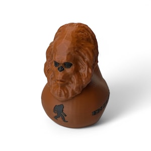 May include: A brown 3D printed figurine of a Bigfoot with a black silhouette of a Bigfoot on the bottom. The figurine is about 3 inches tall.