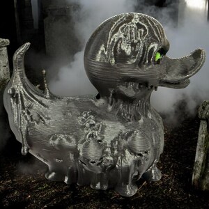 May include: A black rubber duck with a green eye, covered in a textured, dripping, and decaying-looking material. The duck is in a graveyard setting.