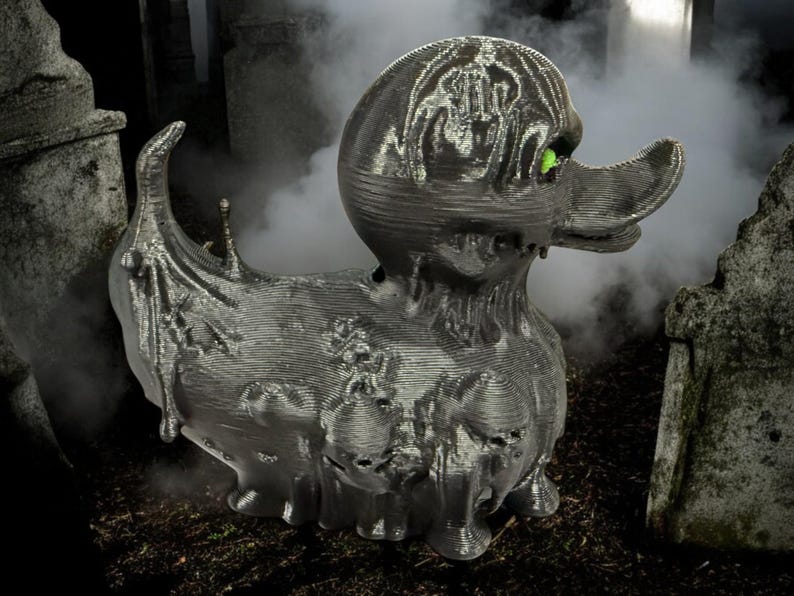 May include: A black rubber duck with a green eye, covered in a textured, dripping, and decaying-looking material. The duck is in a graveyard setting.