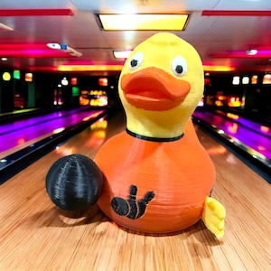 May include: A yellow rubber duck with a bowling ball in its hand, wearing an orange shirt with a bowling pin graphic. The duck is on a wooden bowling lane.