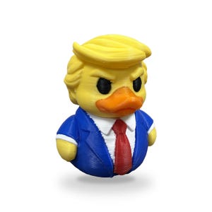 May include: A yellow rubber duck figurine styled with a blonde hairpiece, wearing a blue suit jacket, a white collared shirt, and a red necktie. The duck has an orange beak and black eyes, featuring a textured 3D printed surface finish.