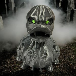 May include: A silver, 3D printed, duck-like creature with glowing green eyes. The creature is standing in a foggy graveyard setting.