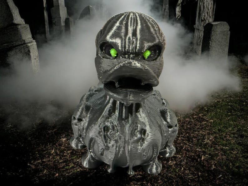 May include: A silver, 3D printed, duck-like creature with glowing green eyes. The creature is standing in a foggy graveyard setting.