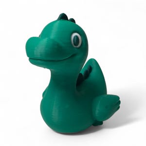 May include: A teal-colored, 3D-printed dinosaur figurine with a friendly expression. The dinosaur has white eyes, a small fin on its back, and a curved tail. The figurine is a solid color and sits on a white background.