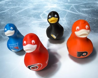 Könnte beinhalten: Vier Gummienten mit unterschiedlichen Farben und Designs. Die Enten sind blau, rot, schwarz und orange, mit Teamlogos auf der Brust. Der Hintergrund ist eine hellblaue, eisähnliche Oberfläche.
