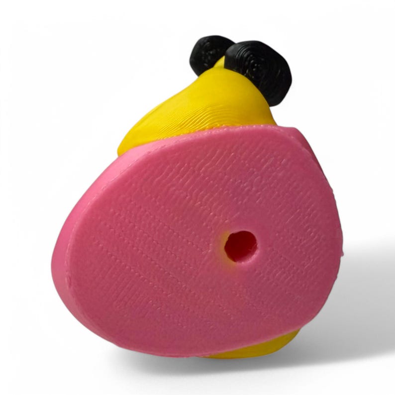 K&ouml;nnte beinhalten: Ein gelbes und rosa 3D-gedrucktes Spielzeug mit einem Loch im Boden. Das Spielzeug hat ein schwarzes und gelbes Design auf der Oberseite.