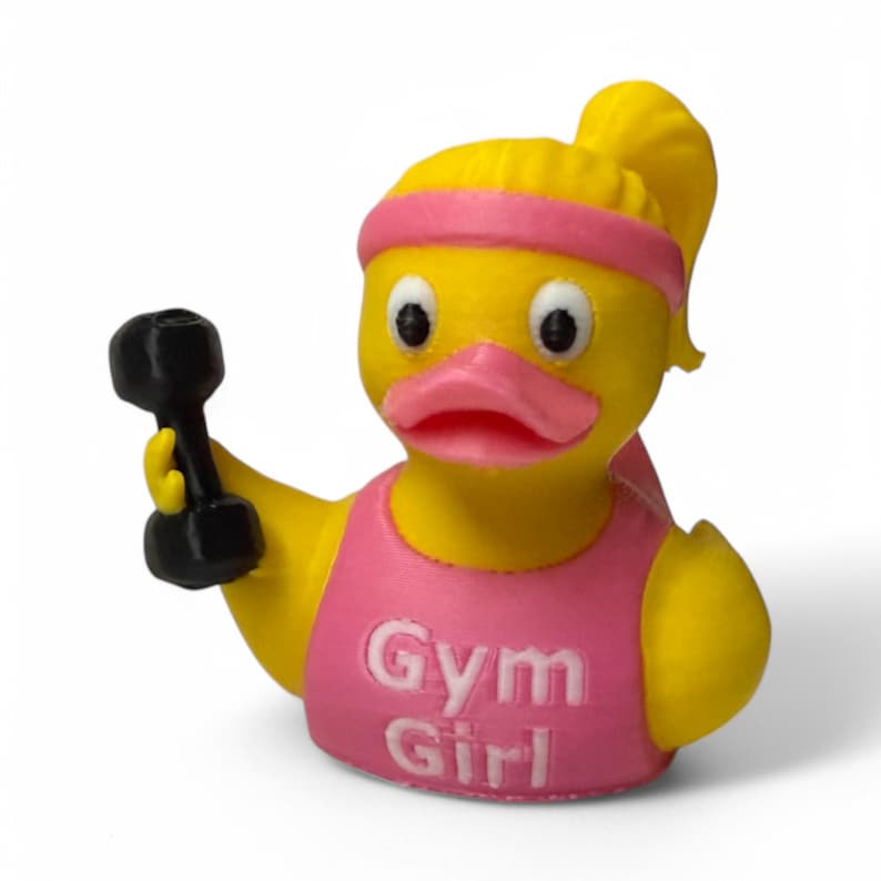 K&ouml;nnte beinhalten: Eine gelbe Gummiente, die ein pinkfarbenes Tanktop mit dem Aufdruck "Gym Girl" tr&auml;gt. Die Ente h&auml;lt eine schwarze Hantel in ihrem rechten Fl&uuml;gel.