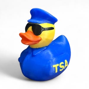 May include: A yellow rubber duck wearing a blue uniform, hat, and sunglasses. The duck has an orange beak and the letters "TSA" in yellow on its chest. The duck is a novelty item.
