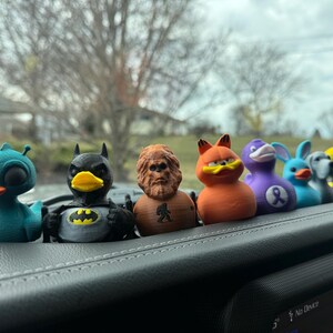May include: A row of seven colorful rubber duck figurines, including a blue duck with an alien-like head, a black and yellow Batman duck, a brown Bigfoot duck, an orange Garfield duck, a purple duck with a ribbon, a blue duck with rabbit ears, and a gray and white duck. The ducks are sitting on a black dashboard.