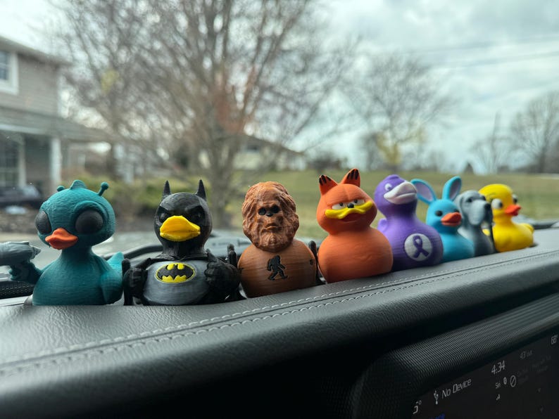 May include: A row of seven colorful rubber duck figurines, including a blue duck with an alien-like head, a black and yellow Batman duck, a brown Bigfoot duck, an orange Garfield duck, a purple duck with a ribbon, a blue duck with rabbit ears, and a gray and white duck. The ducks are sitting on a black dashboard.