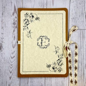 May include: A beige journal with a brown border and a decorative floral design. The journal features the letter "E" in a wreath and is tied with a ribbon printed with brown insects.