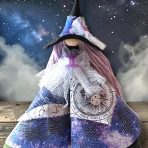 May include: A handmade witch doll with a black hat, purple hair, and a white dress. The doll's dress is decorated with a celestial design, including constellations and a zodiac wheel. The background features a starry night sky.