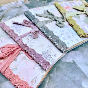 May include: Three journals with floral patterned covers, each featuring a decorative lace spine trim and a matching fabric ribbon tie. The journals are arranged in a row, showcasing covers in shades of pink, sage green, and mustard yellow.